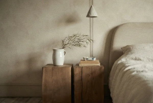 Minimalist bedroom scene with wooden side tables, a white ceramic pitcher holding branches, books, and a soft bed with white bedding.