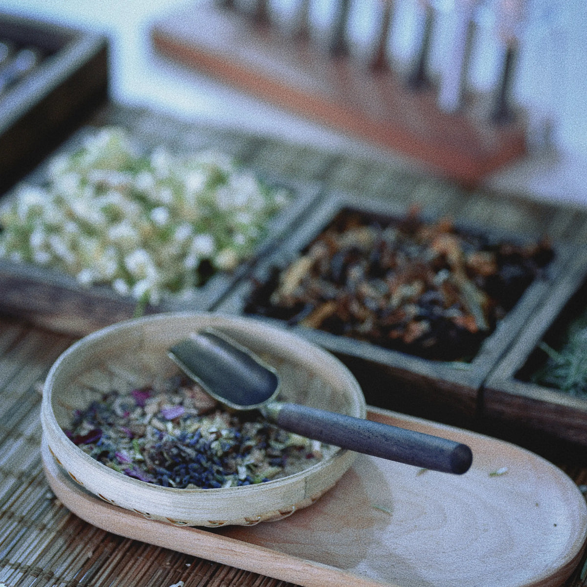 A close-up of loose herbal tea or spices in a small bamboo bowl with a dark scoop, set on a wooden tray with other dried ingredients in the background.