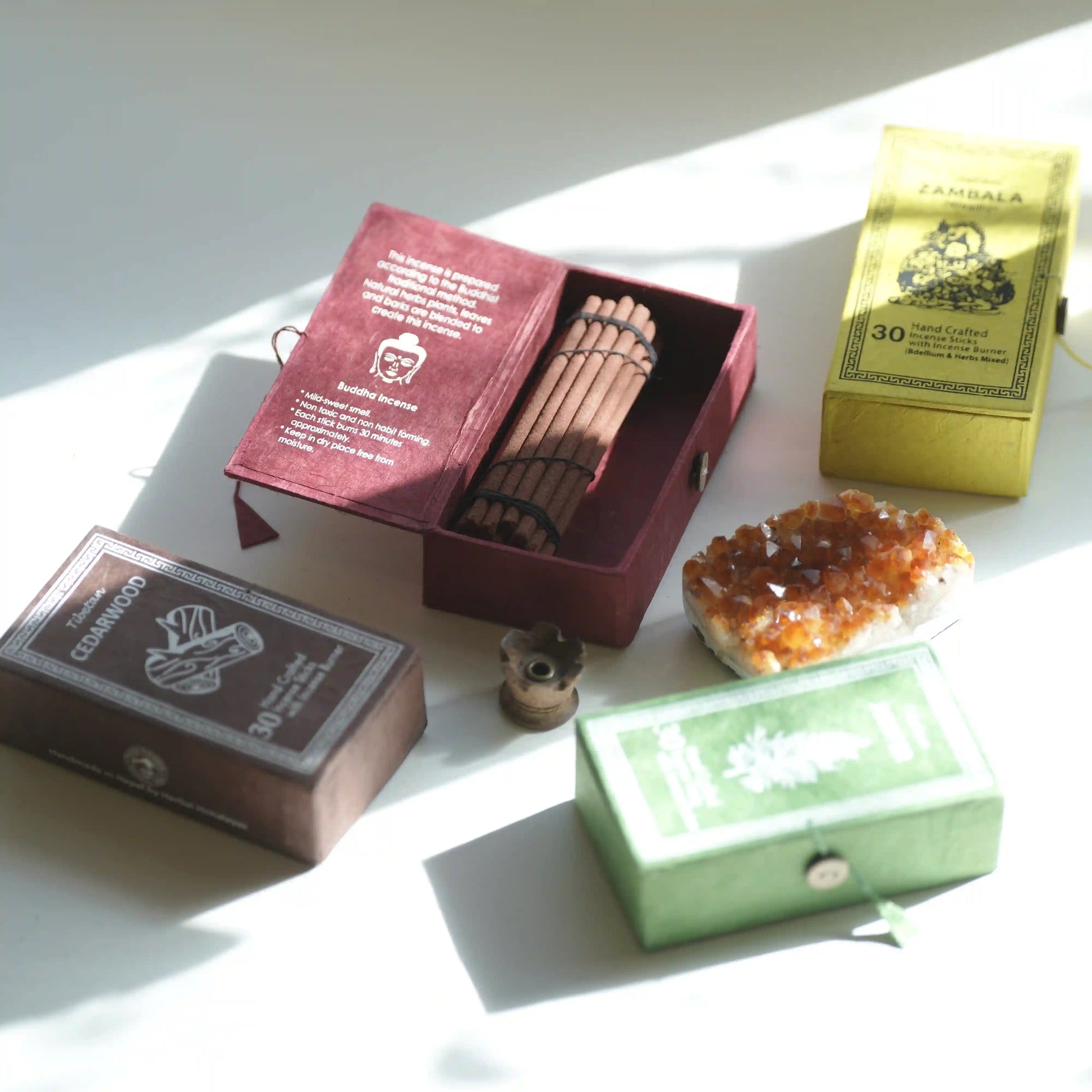 A styled flat-lay of several miniature traditional incense burners and their matching boxes on a white surface with strong diagonal sunlight. The set includes a dark brown box labeled 'CANDUFA', a dark red open box containing a bamboo incense holder, a yellow box labeled 'JAMBALA 30 CIGS', a green box, and a natural orange crystal cluster.