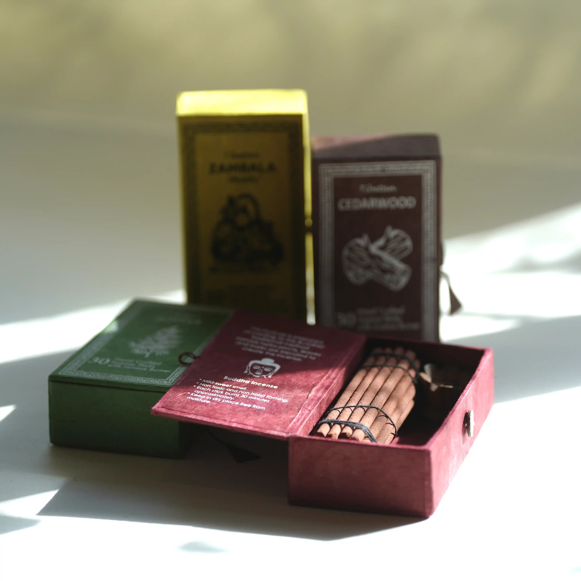 Four paper box instruments arranged on a white surface in bright natural light. An open burgundy box in front holds a set of stringed Tibetan Incense , with two upright boxes behind them in yellow and dark brown, and one green box lying on its side.