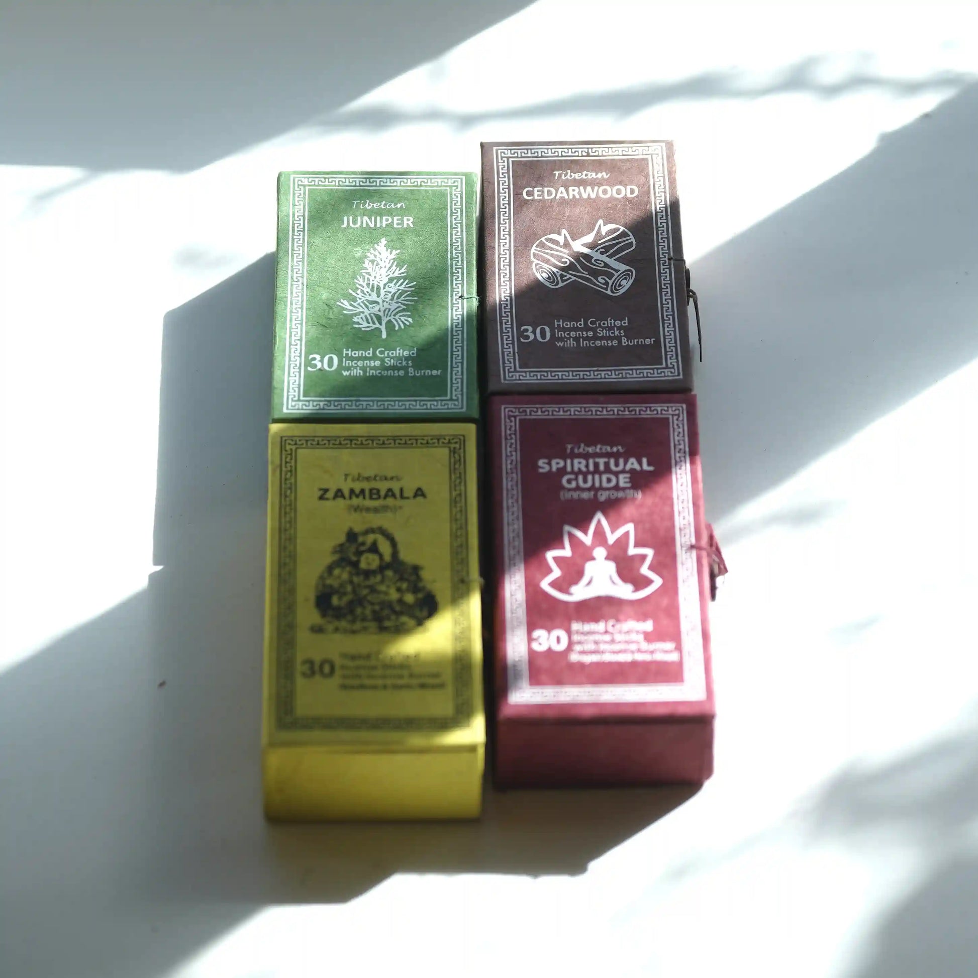 Four boxed Tibetan prayer flags arranged in a 2x2 grid on a white surface with strong natural shadows. The boxes are labeled Tibetan Incense Juniper, Cedarwood, Zambala, and Spiritual Guide, with colors green, brown, yellow, and burgundy.