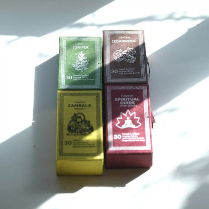 Four boxed Tibetan prayer flags arranged in a 2x2 grid on a white surface with strong natural shadows. The boxes are labeled Tibetan Incense Juniper, Cedarwood, Zambala, and Spiritual Guide, with colors green, brown, yellow, and burgundy.