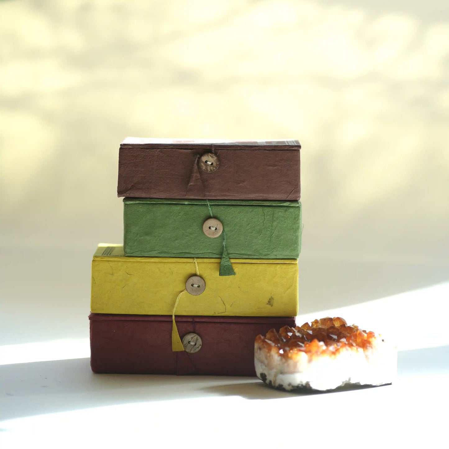 A stack of four Tibetan Incense  boxes  arranged vertically on a light surface. The boxes are brown, green, yellow, and dark red/burgundy.