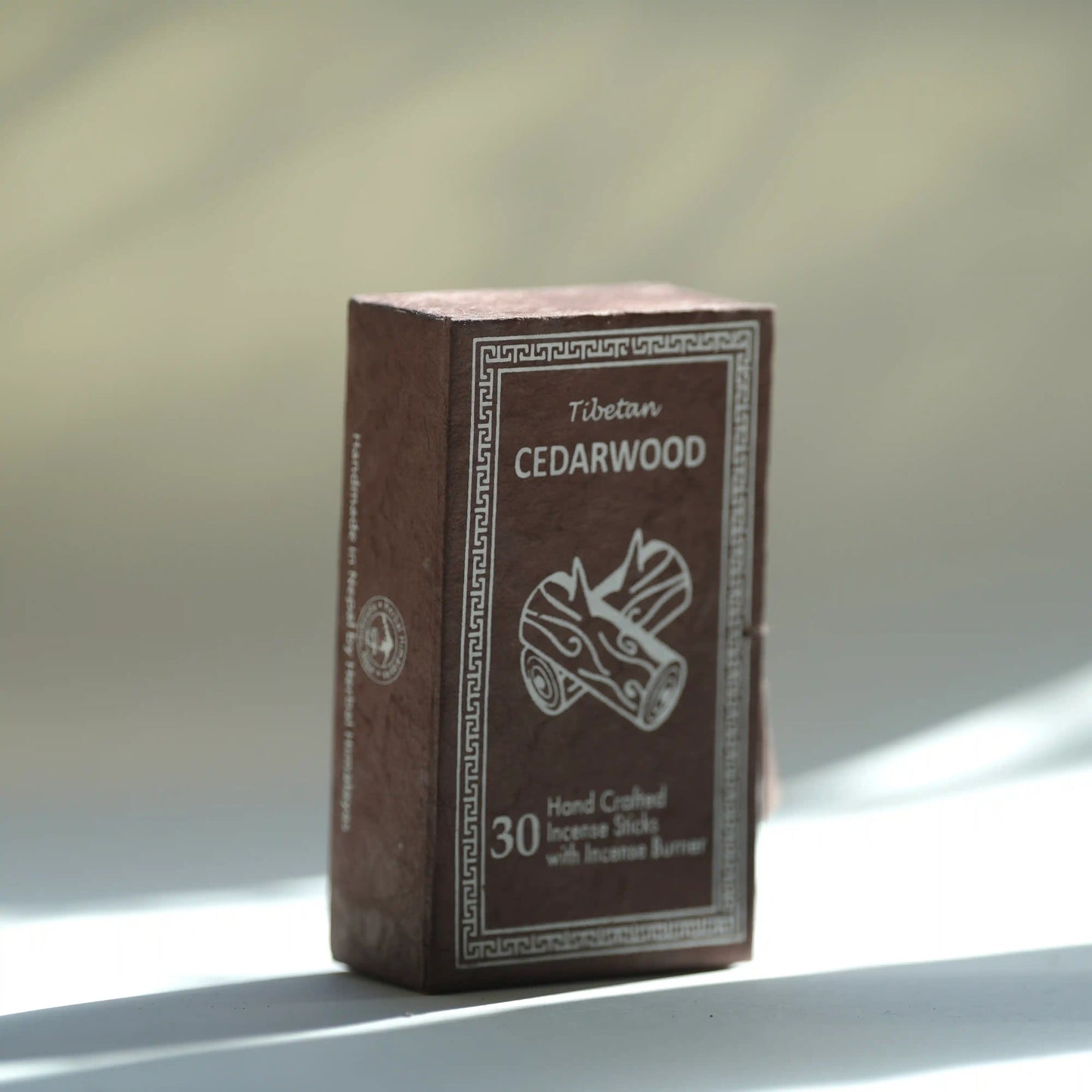 A box of Tibetan cedarwood incense sticks standing upright on a light surface with soft natural shadows. The box is dark brown with white text and an illustration of a incense burner and logs.