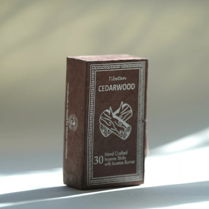 A box of Tibetan cedarwood incense sticks standing upright on a light surface with soft natural shadows. The box is dark brown with white text and an illustration of a incense burner and logs.