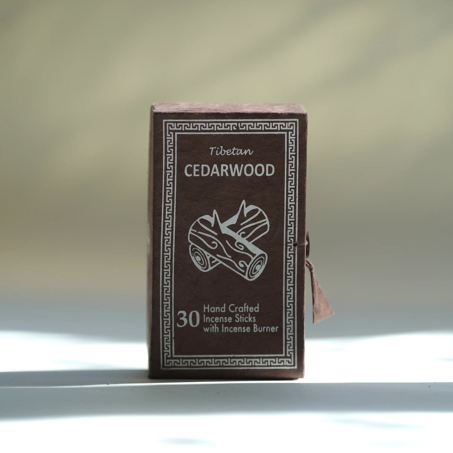 A small rectangular box of Tibetan cedarwood incense sticks stands upright on a white surface against a softly blurred light gray background. The box is dark brown with white printed text and border detailing, including the words 'Tibetan CEDARWOOD' and '30 Hand Crafted Incense Sticks with Incense Burner.