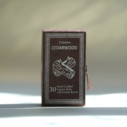 A small rectangular box of Tibetan cedarwood incense sticks stands upright on a white surface against a softly blurred light gray background. The box is dark brown with white printed text and border detailing, including the words 'Tibetan CEDARWOOD' and '30 Hand Crafted Incense Sticks with Incense Burner.