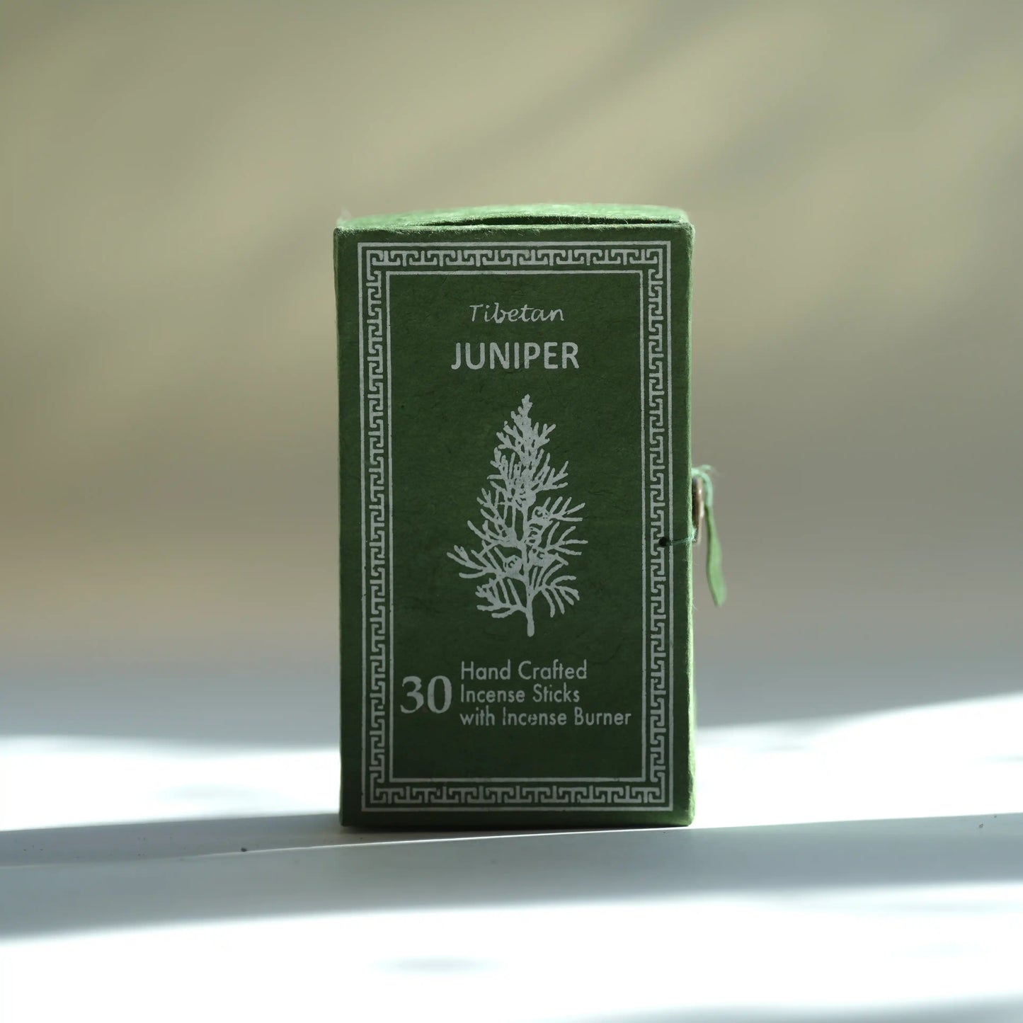 A dark green box of Tibetean Juniper handcrafted incense sticks with an incense burner, featuring a white tree illustration and Greek key border, set against a softly blurred beige background with light shadows.