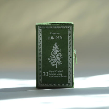 A dark green box of Tibetean Juniper handcrafted incense sticks with an incense burner, featuring a white tree illustration and Greek key border, set against a softly blurred beige background with light shadows.