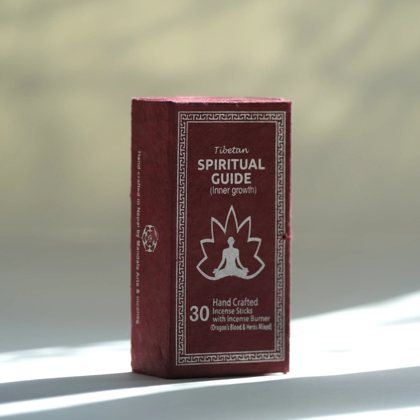 A boxed Tibetan Spiritual Guide incense stick set standing upright on a light surface with a softly blurred warm-toned background. The box is dark red/burgundy with white text and a white meditating figure icon.