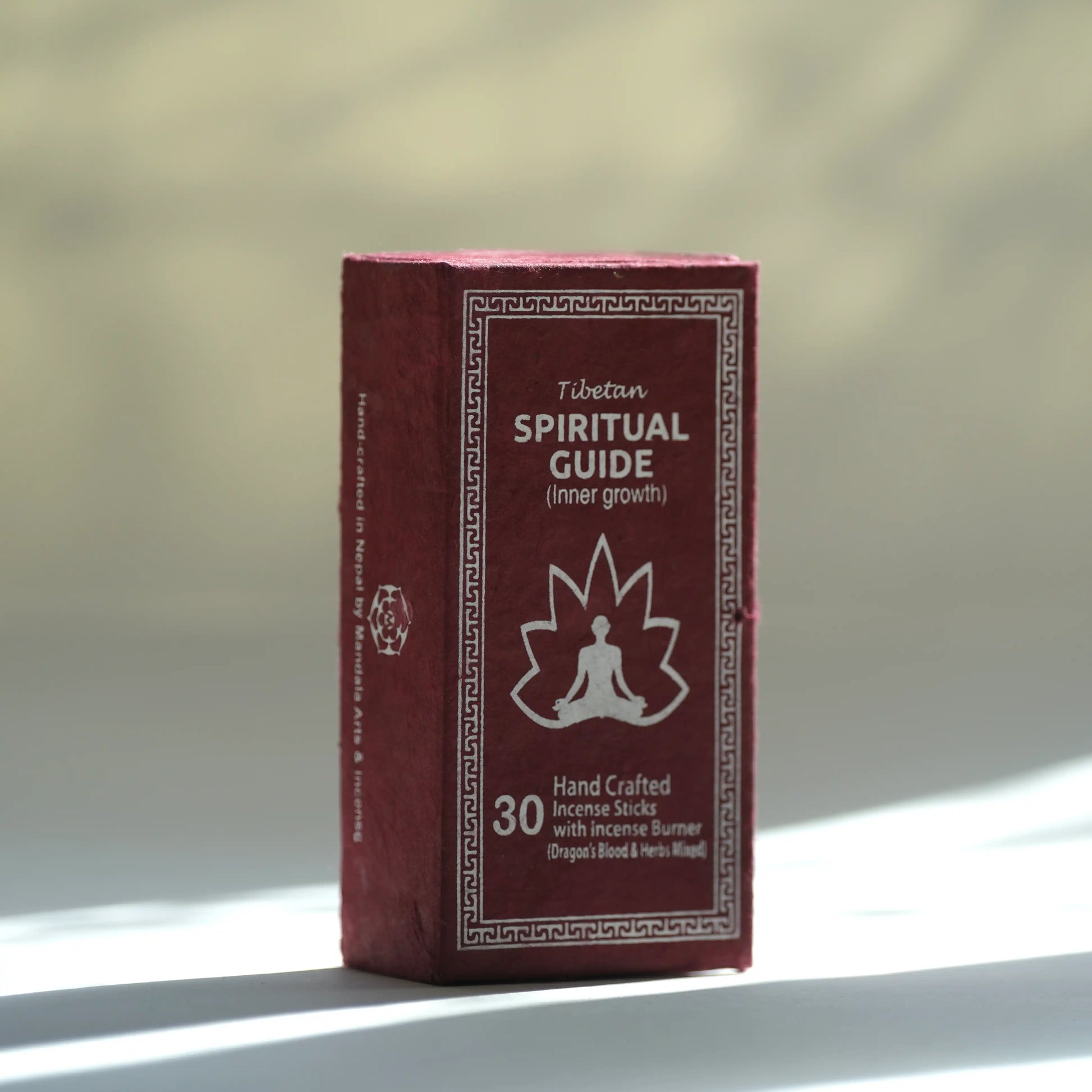 A boxed Tibetan Spiritual Guide incense stick set standing upright on a light surface with a softly blurred warm-toned background. The box is dark red/burgundy with white text and a white meditating figure icon.