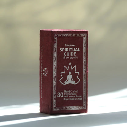 A boxed Tibetan Spiritual Guide incense stick set standing upright on a light surface with a softly blurred warm-toned background. The box is dark red/burgundy with white text and a white meditating figure icon.