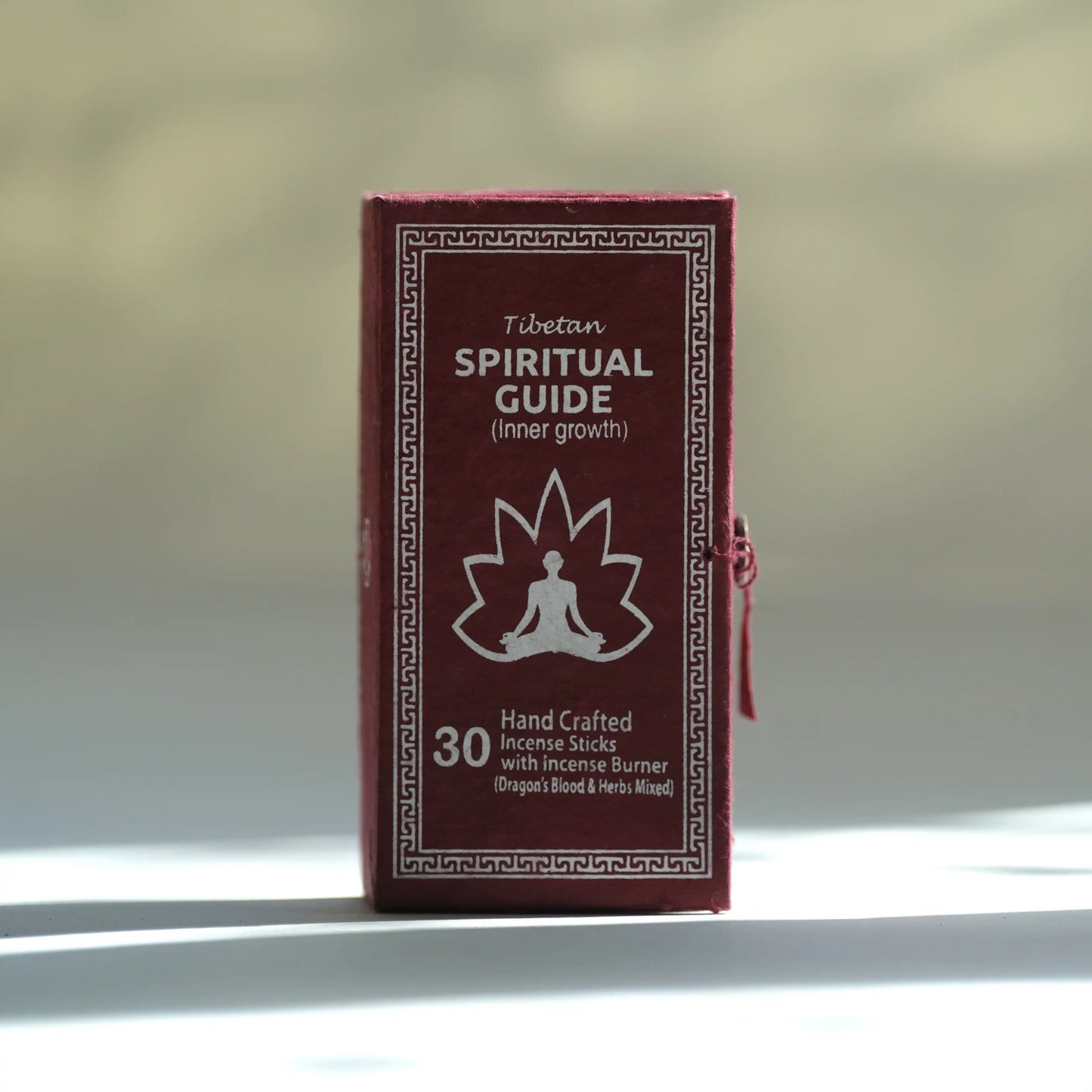 A small rectangular box labeled 'Tibetan Spiritual Guide (inner growth)' with a white meditating figure and maple leaf graphic on a dark red front, standing upright against a softly blurred beige background with bright sunlight and shadows.