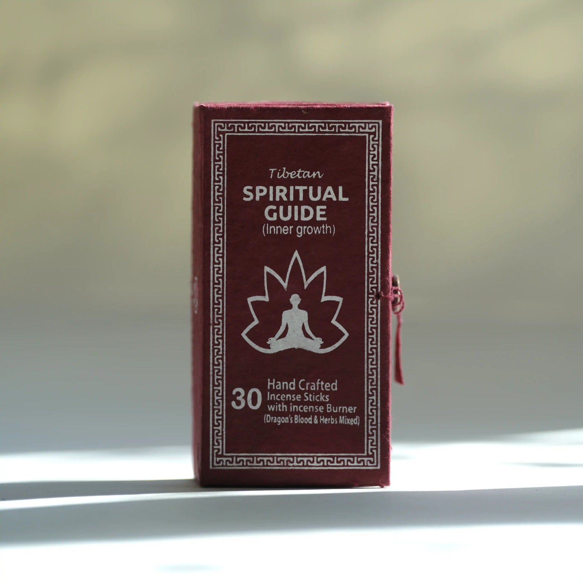 A small rectangular box labeled 'Tibetan Spiritual Guide (inner growth)' with a white meditating figure and maple leaf graphic on a dark red front, standing upright against a softly blurred beige background with bright sunlight and shadows.
