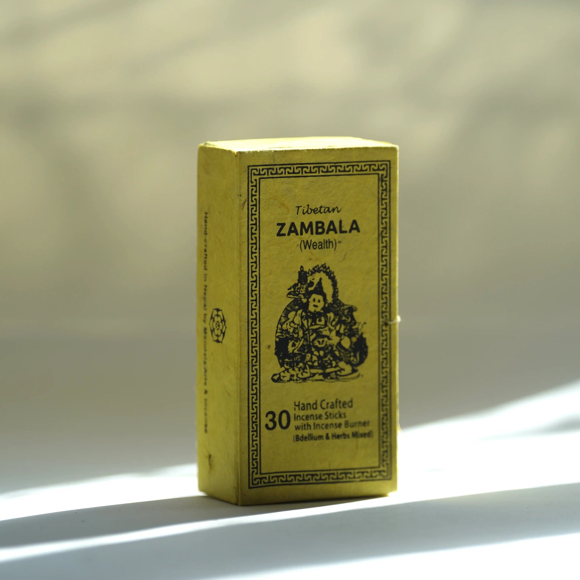 A yellow box of Tibetan Zambala (wealth) handcrafted incense sticks, standing on a white surface with soft, dappled sunlight and shadows.