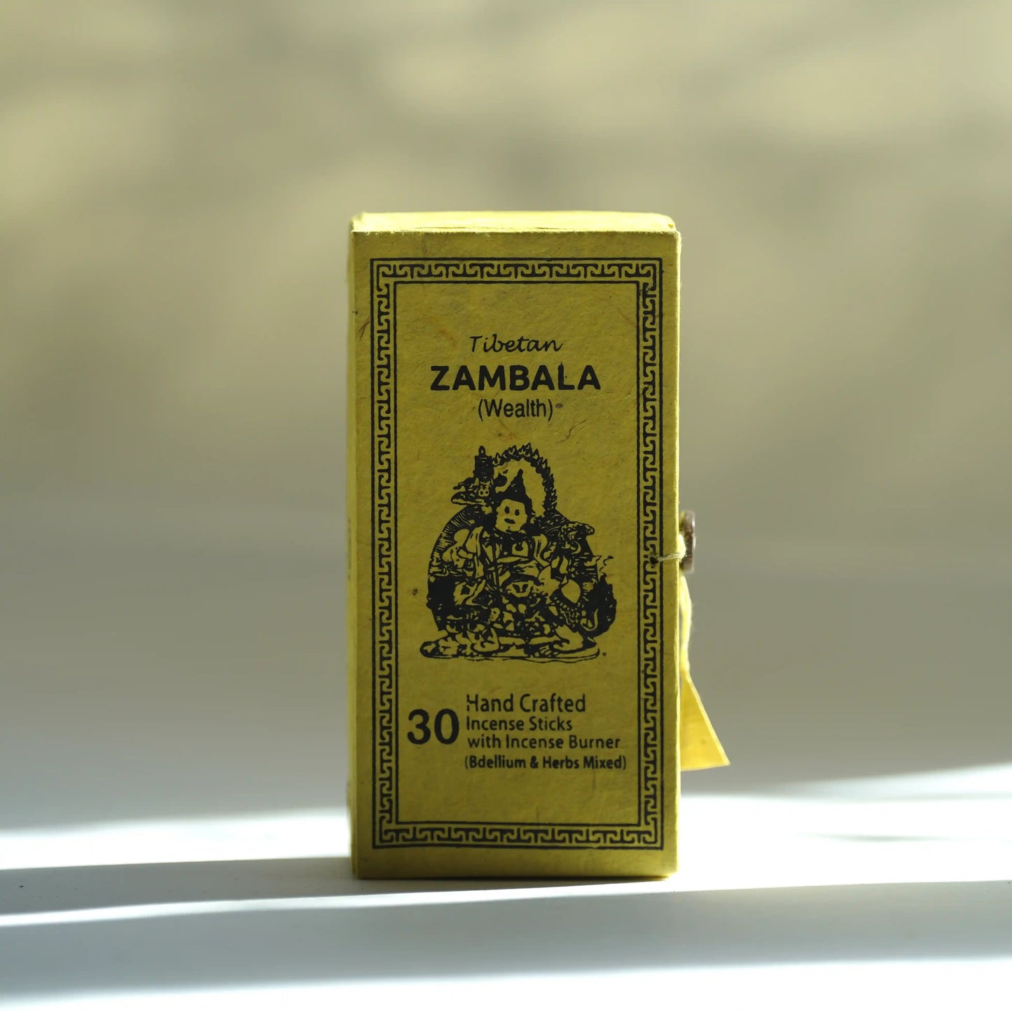 A small rectangular yellow box of Tibetan Zambala incense sticks stands upright on a white surface with soft sunlight and shadows. The front shows black text and an illustration, with a small yellow ribbon tied to the side.