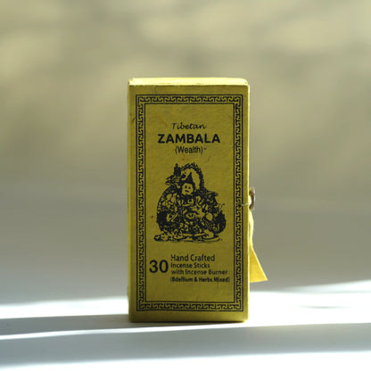 A small rectangular yellow box of Tibetan Zambala incense sticks stands upright on a white surface with soft sunlight and shadows. The front shows black text and an illustration, with a small yellow ribbon tied to the side.