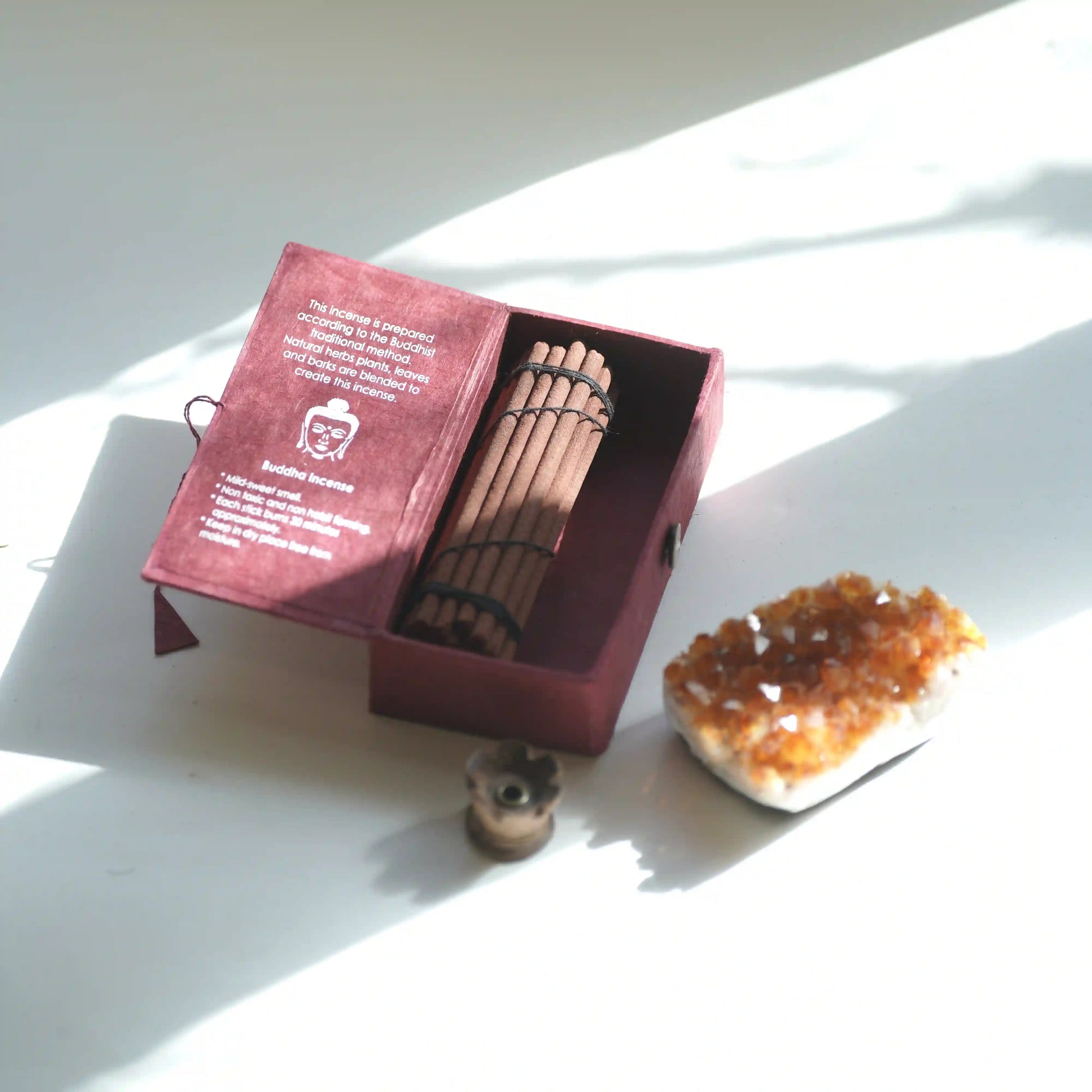 An open burgundy presentation box containing a set of Tibetan incense sticks, with printed text visible on the inside lid. A small incense burner sits in front, and a crystal cluster rests beside the box on a white surface in bright natural light.