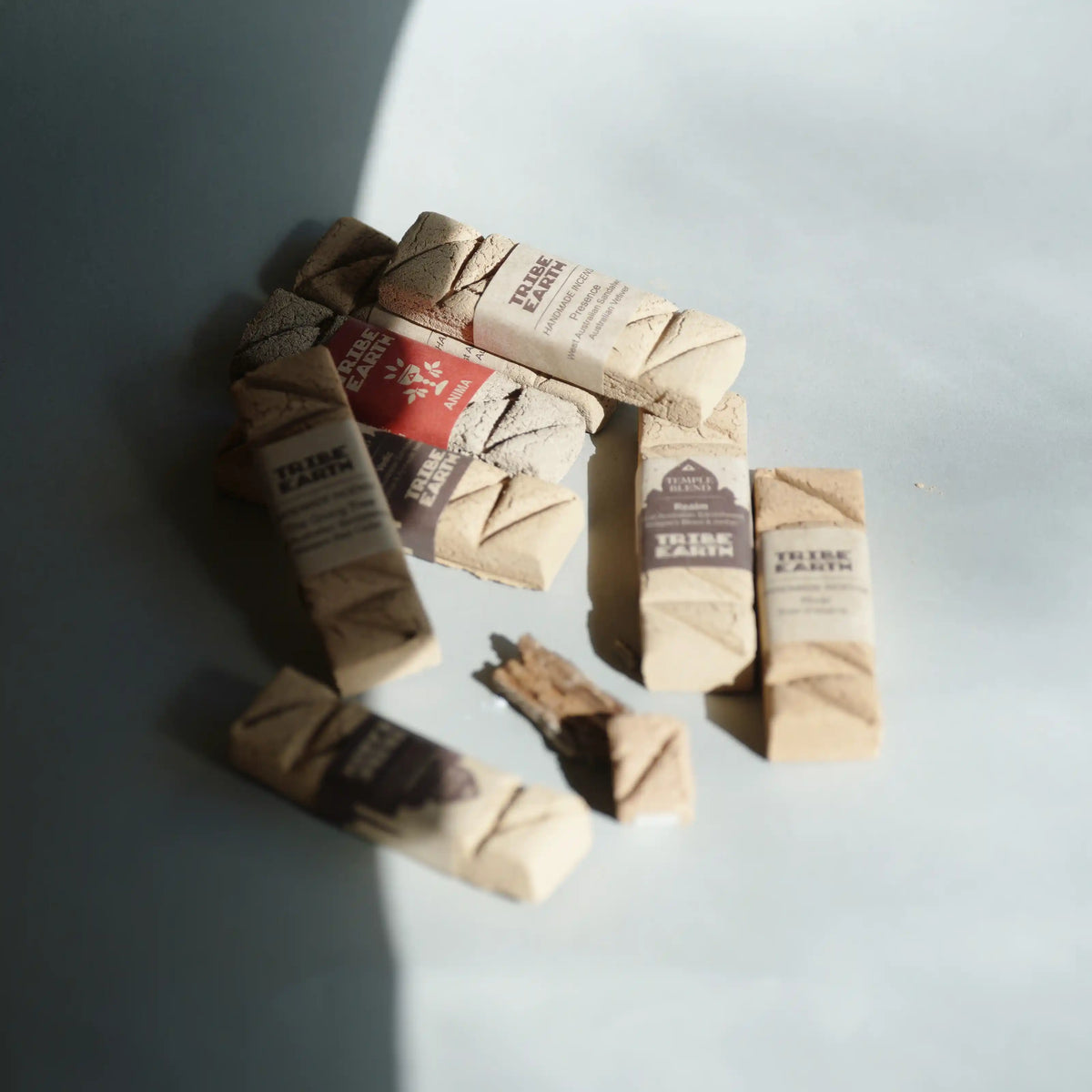 Several small wrapped Trie earth incense are arranged on a pale blue-gray surface with strong diagonal sunlight and shadows.