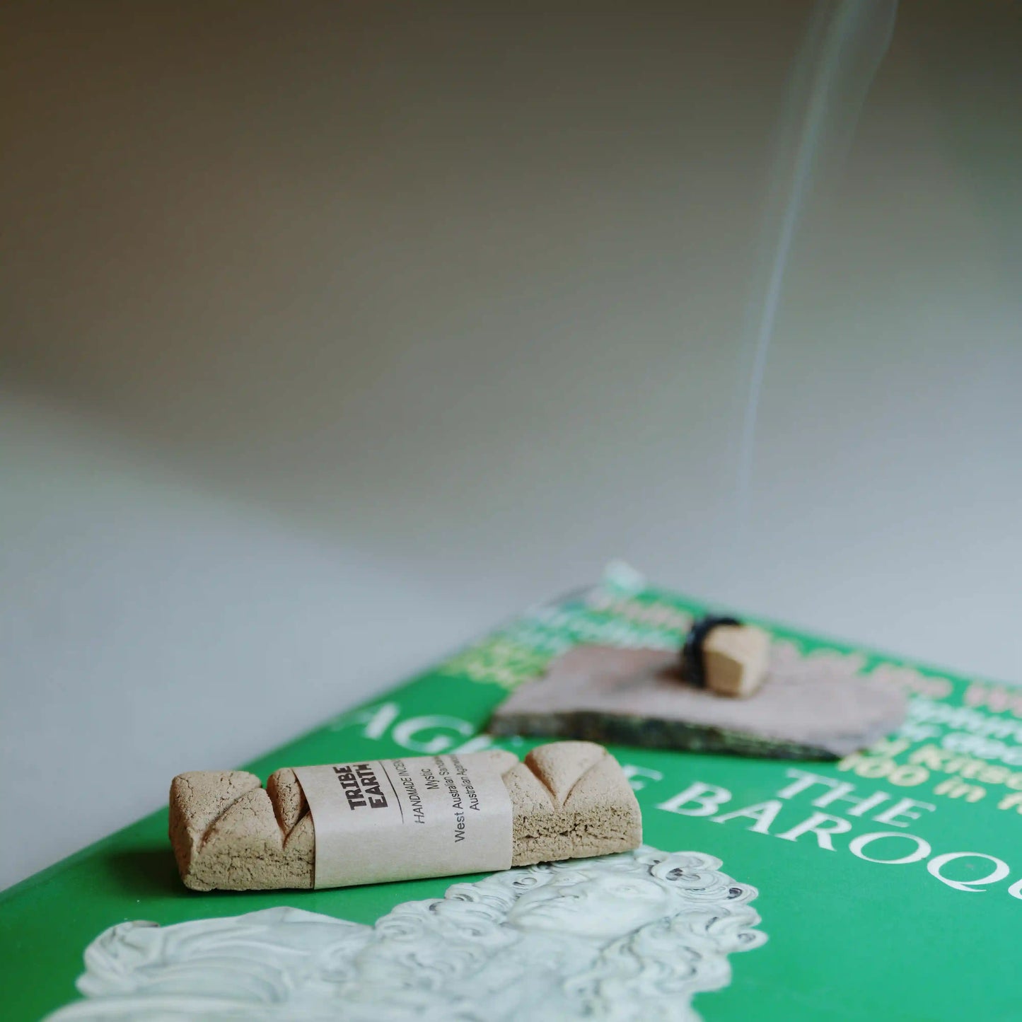 A rolled bundle of incense sticks wrapped in a paper band sits on a green book, with a second incense stick burning and smoke rising in the background.