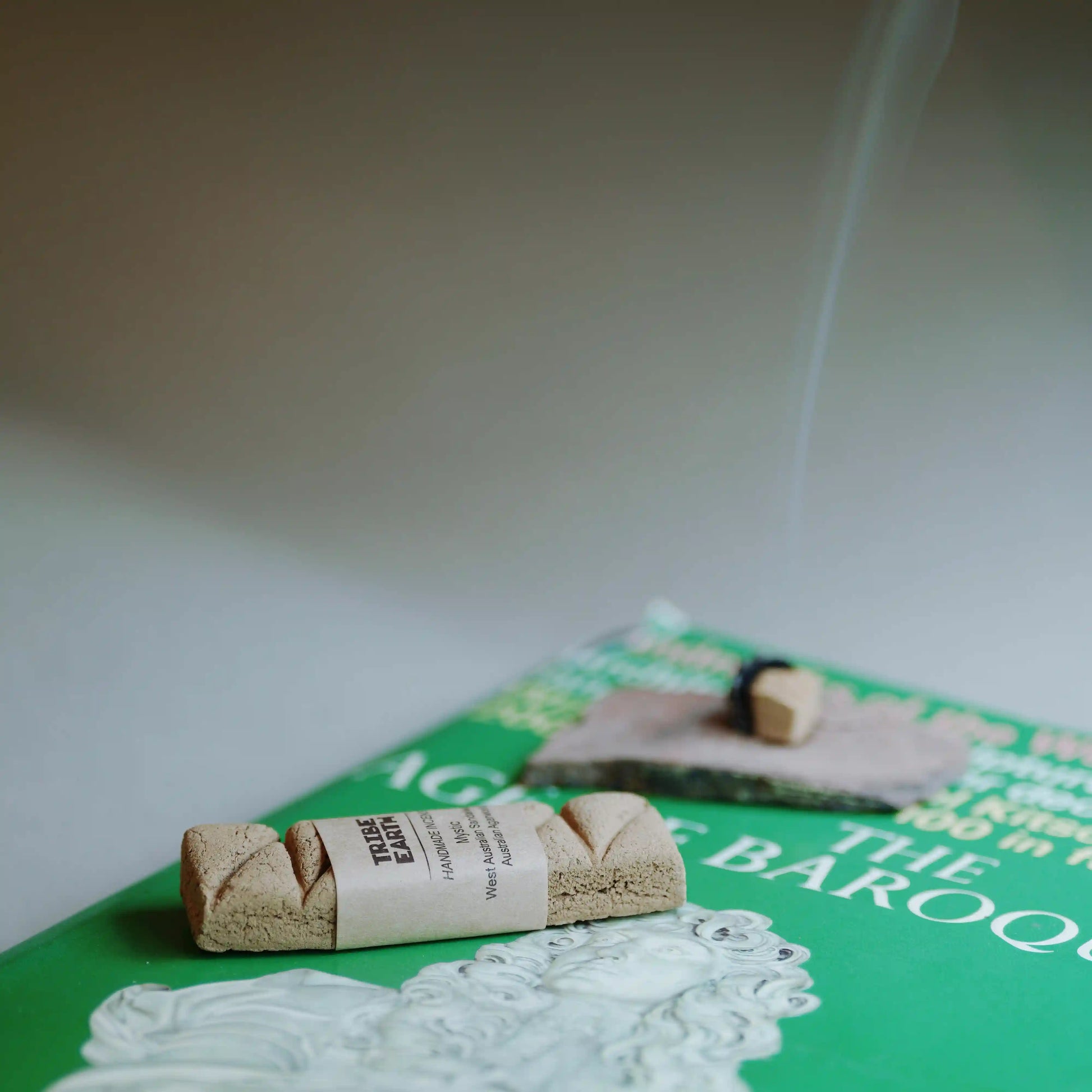 A rolled bundle of incense sticks wrapped in a paper band sits on a green book, with a second incense stick burning and smoke rising in the background.