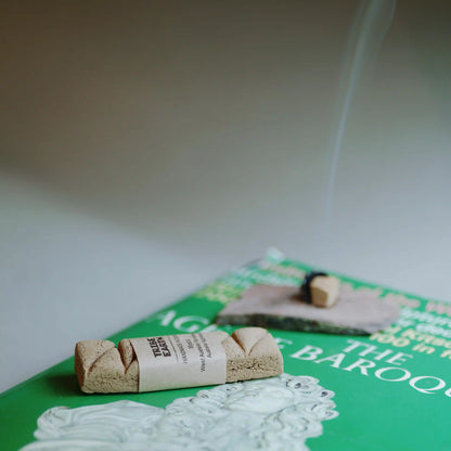A rolled bundle of incense sticks wrapped in a paper band sits on a green book, with a second incense stick burning and smoke rising in the background.