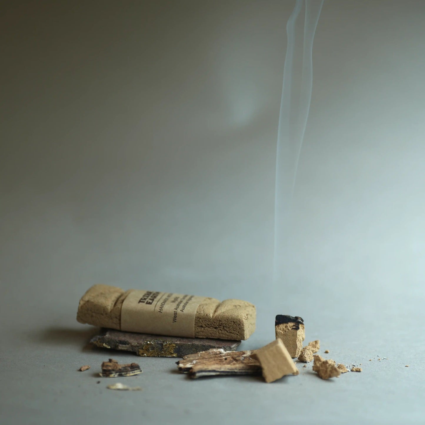 A bundle of natural cork incense sticks lies on a gray surface with a few broken pieces scattered around. A thin white smoke curl rises into the background against a neutral gray studio backdrop.