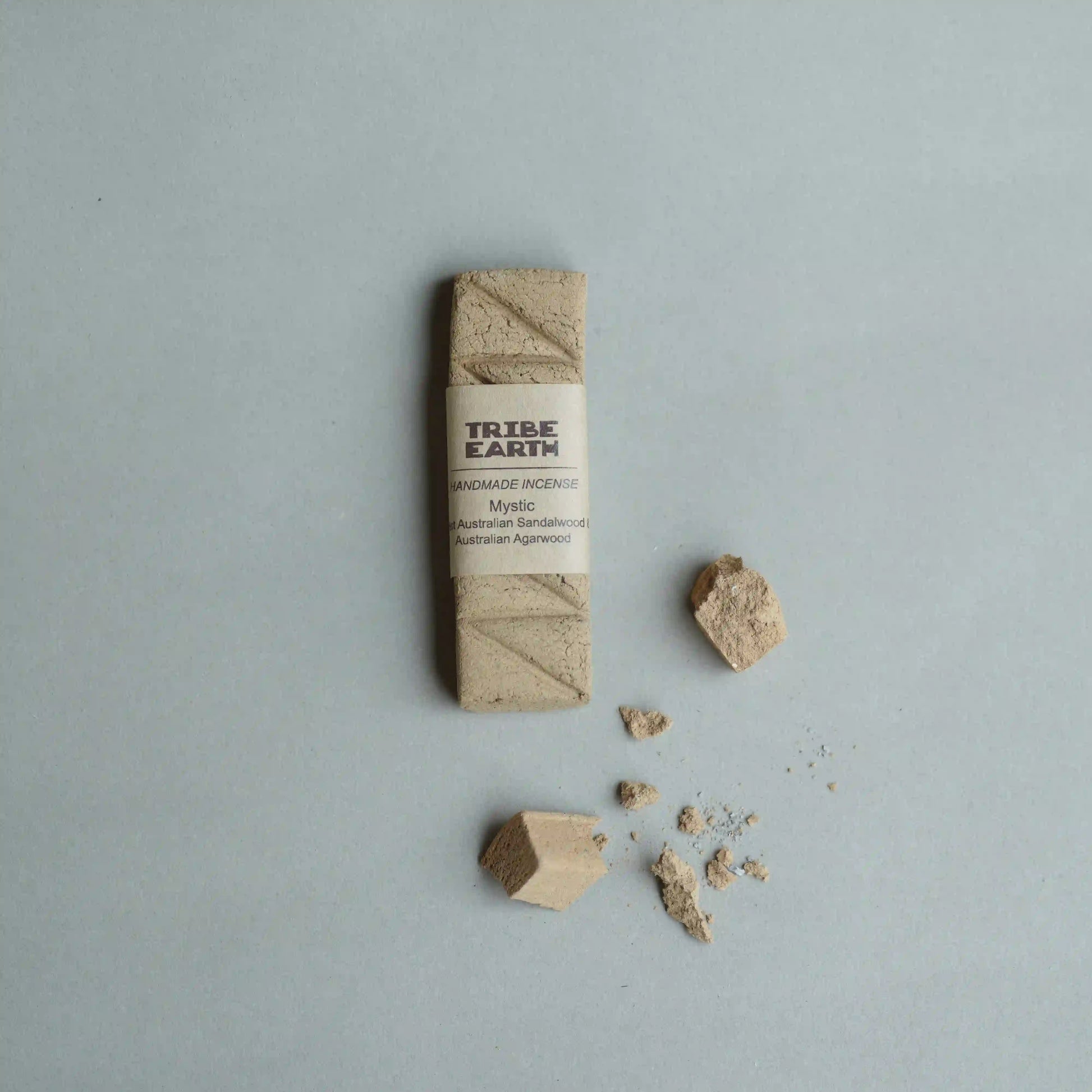 A handcrafted incense wrapped in a paper band labeled 'TRIBE EARTH HANDMADE INCENSE' on a light gray background, with small pieces of the natural wood and powder scattered nearby.