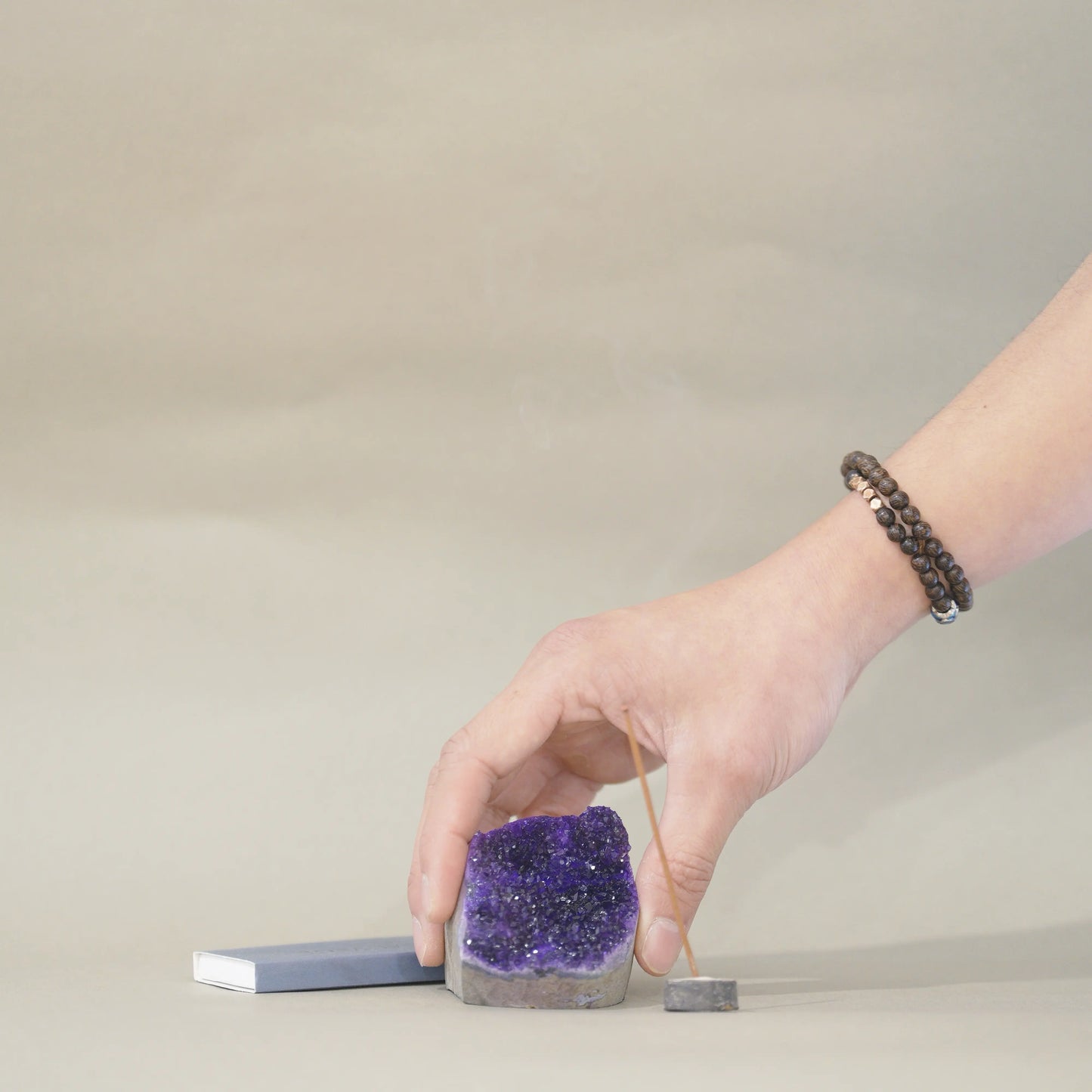 A hand wearing a dark beaded bracelet places a small incense stick into a square gray stone holder next to a purple amethyst geode and a blue incense box on a neutral beige background.