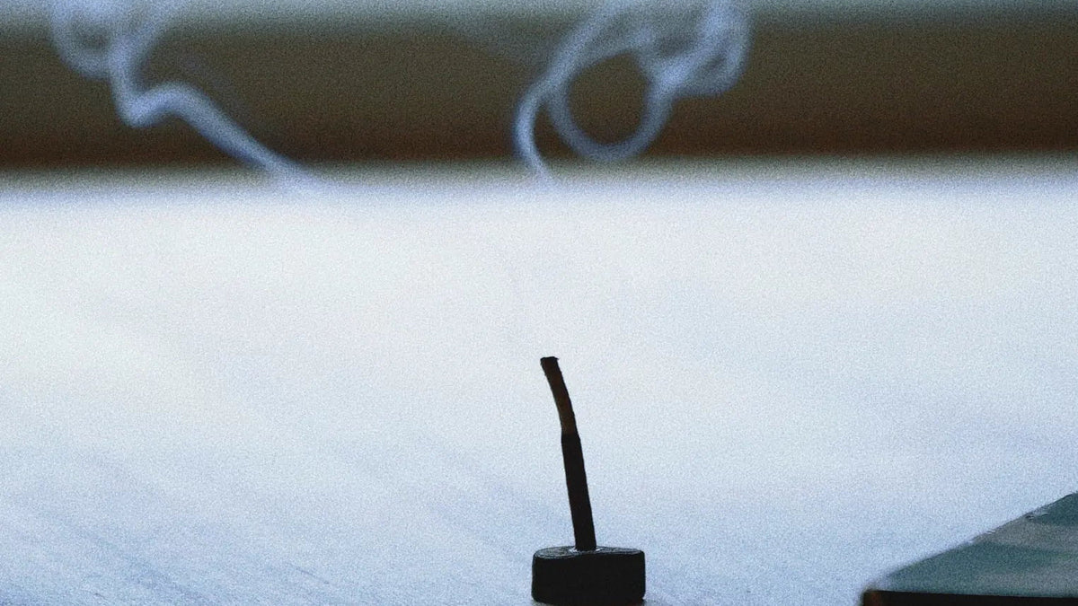 A small black incense holder with a single lit incense stick, with white smoke curling upward on a light wooden surface in front of a blurred green window background.