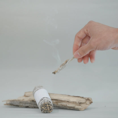 A hand holds a small smudging bundle above another resting on driftwood against a clean light gray background.