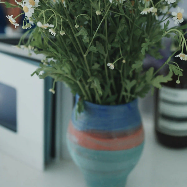A close-up of a colorful ceramic vase holding green stems with small white daisy-like flowers on a white surface.