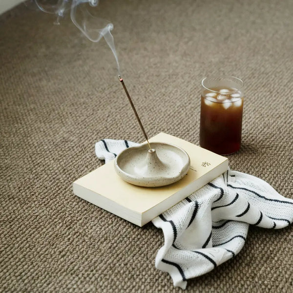 Incense stick burning in a ceramic holder on a closed book with a striped cloth and a glass of iced coffee on carpet.