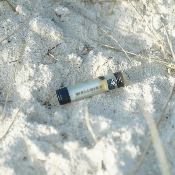 Small cylindrical WellHike torch lying on sandy ground with sparse dry grass.