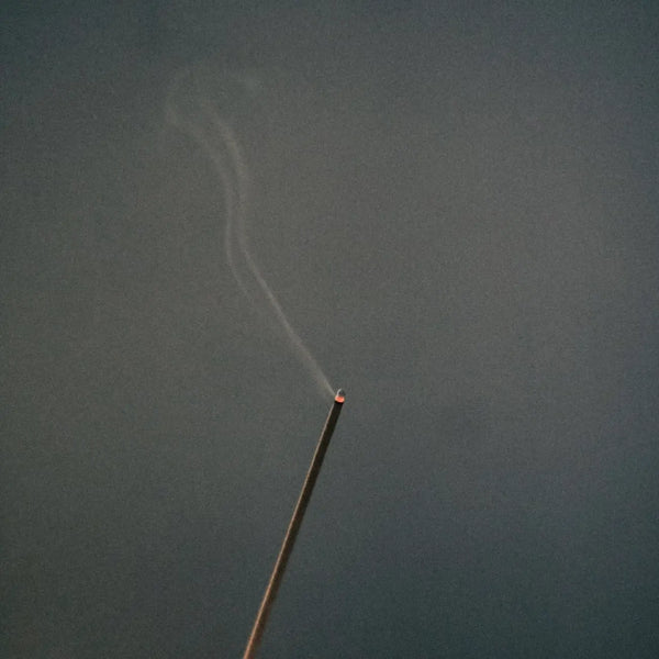 Thin incense stick with a glowing tip emitting a light trail of smoke against a dark gray background.