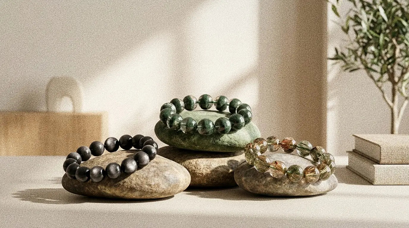 A curated collection of genuine gemstone bead bracelets for energy, protection, and balance. The minimalist display showcases a black silver sheen obsidian bracelet, a green phantom quartz bracelet, and a rutilated quartz crystal bracelet on natural earthy stones.