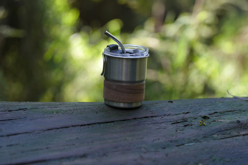 Stainless steel reusable coffee cup with wooden sleeve resting on a wooden surface in a natural outdoor setting