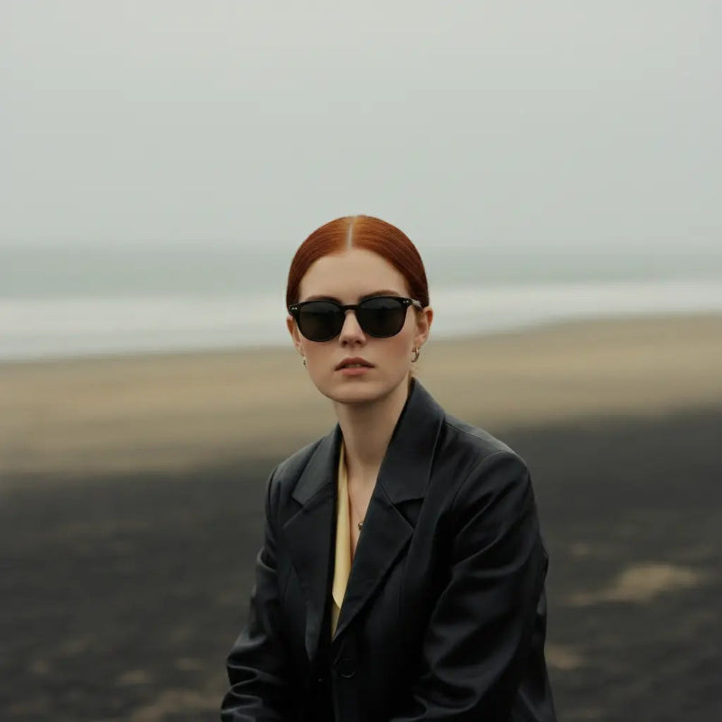 A person with red hair wears a black blazer, seated against a moody beach backdrop with muted colors and soft waves in the distance.